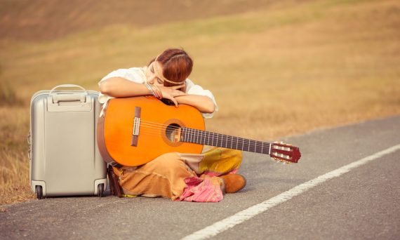 Young hippie woman with guitar and backpack sitting on a countryside road